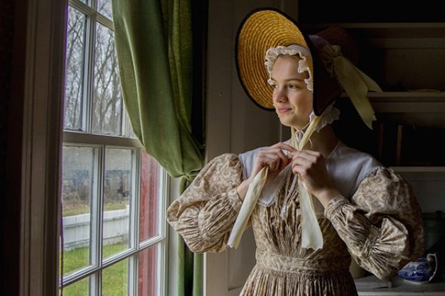 A pleasant looking young lady wearing period attire a really big bonnet-style hat looking out of a window in winter