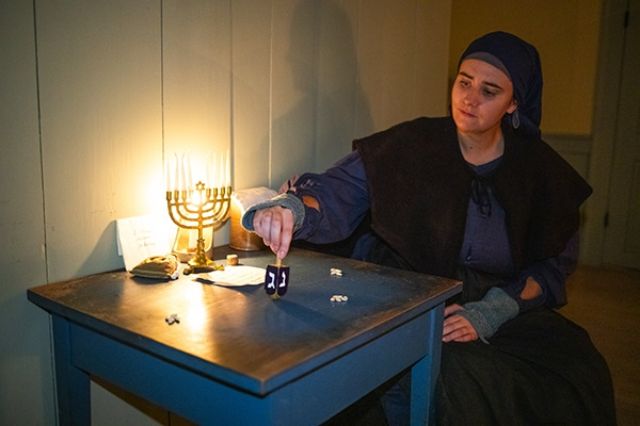 A lady wearing some traditional looking clothing sitting at a square worn table with a menorah and a really big sweet looking dreidel