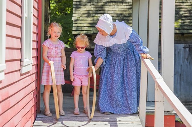 A lady in period attire teaching two young girls how to play the hoop game apparently everyone loves the hoop game