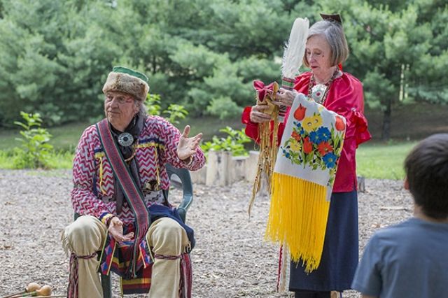 An older man in a pretty authentic looking prairie outfit excellent hat alongside a woman with some artifacts - they appear to be telling a story to a group of children