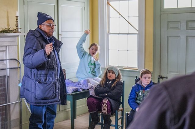 A man in a big coat and beanie warm hat is telling a story to some school-aged children who look cold and one is trying to distract him with a question that may or may not be relevant.