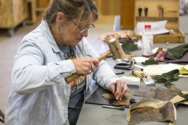 Lady with a Fred Flintstone style mallet pounding a hole in a sheet of leather with an awl she may be about to smash a finger.