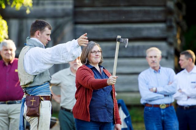 A lady learning the useful skill of axe throwing while men watch - everyone seems calm.